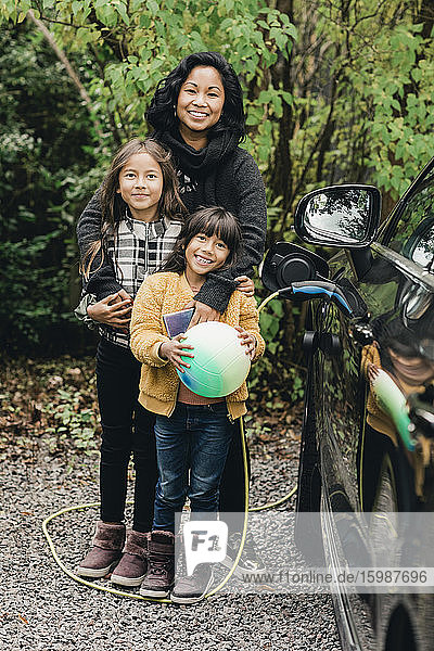 Porträt einer lächelnden Frau mit Töchtern in einem Elektroauto an einer Ladestation