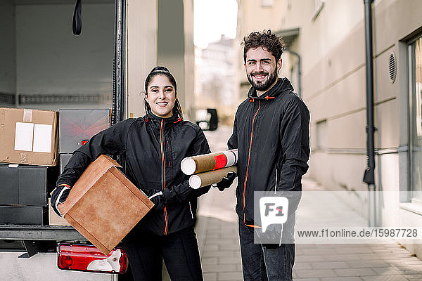 Portrait of confident delivery man and woman with packages standing against truck in city