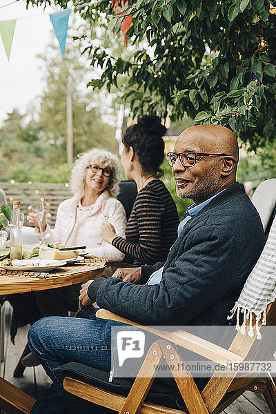 Lächelnder kahlköpfiger älterer Mann sitzt auf Stuhl und genießt Dinnerparty von Freunden im Hinterhof