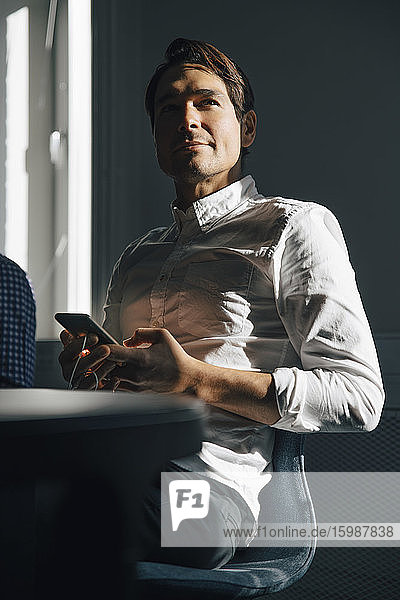 Selbstbewusster Geschäftsmann sitzt mit Smartphone am Konferenztisch im Sitzungssaal