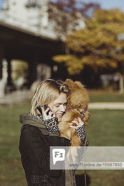 Smiling blond woman talking on mobile phone while carrying Pomeranian at park on sunny day