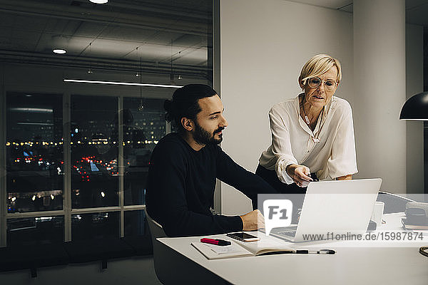 Männliche und weibliche Fachkräfte planen ihre Strategie  während sie über einen Laptop in einem beleuchteten Büro diskutieren