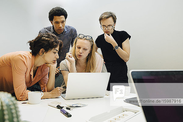 Multi-ethnic colleagues planning strategy while looking at laptop in illuminated office board room
