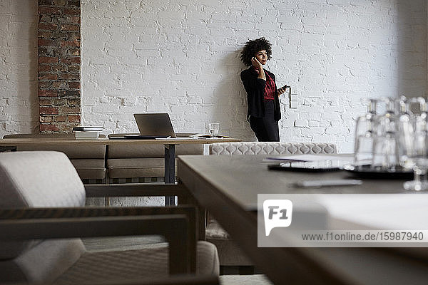 Selbstbewusste junge Anwältin telefoniert mit dem Handy  während sie sich im Büro an eine weiße Wand lehnt