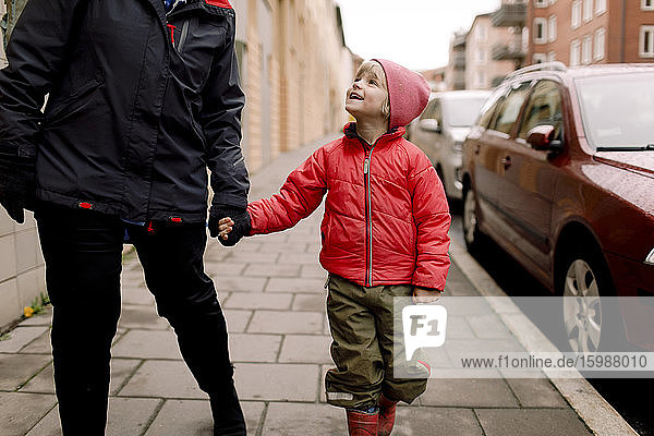 Lächelnder Enkel in warmer Kleidung hält die Hände der Großmutter beim Gehen auf dem Fußweg