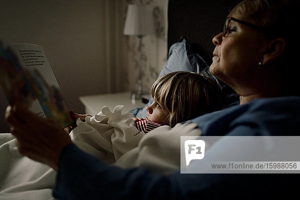 Senior woman reading story book for grandson on bed at home