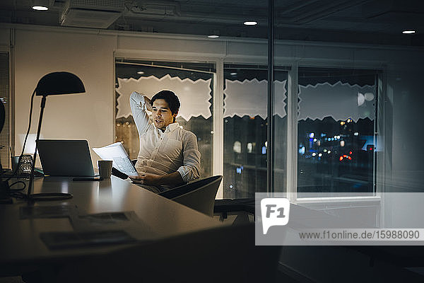 Confident male entrepreneur reading document while sitting in dark office at night