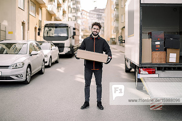 Portrait of confident young delivery man with box leaning on truck
