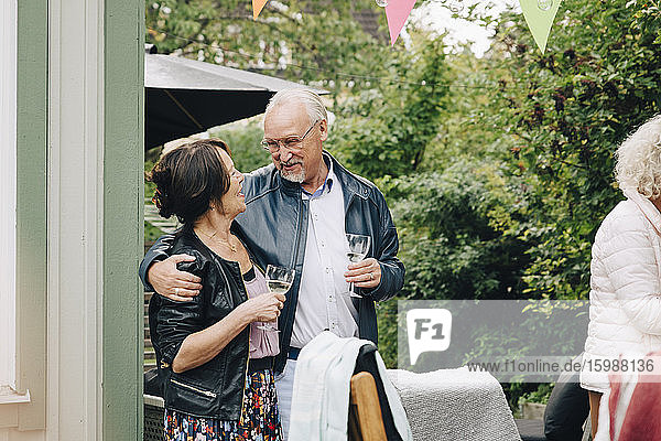 Smiling senior friends standing with arm around enjoying garden party at back yard