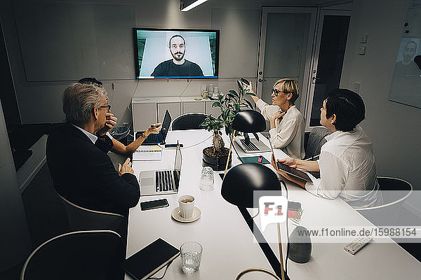 Männliche und weibliche Kollegen diskutieren mit einem Geschäftsmann per Videoanruf im Sitzungssaal während einer Sitzung spät in der Nacht