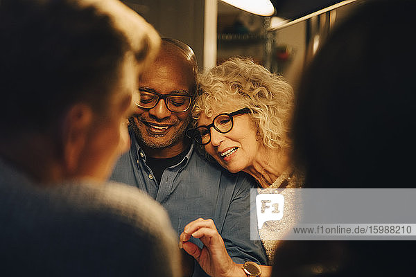 Lächelnder Senior teilt Smartphone mit Frau  während er am beleuchteten Esstisch sitzt