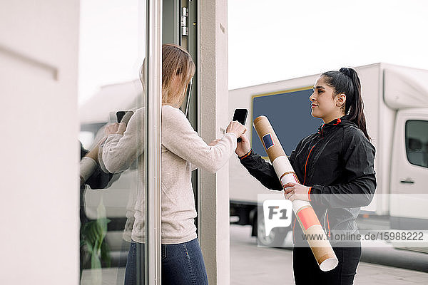 Female customer signing on smart phone while receiving package from delivery woman