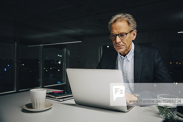 Selbstbewusster Senior-Geschäftsmann mit Laptop am beleuchteten Schreibtisch im Kreativbüro