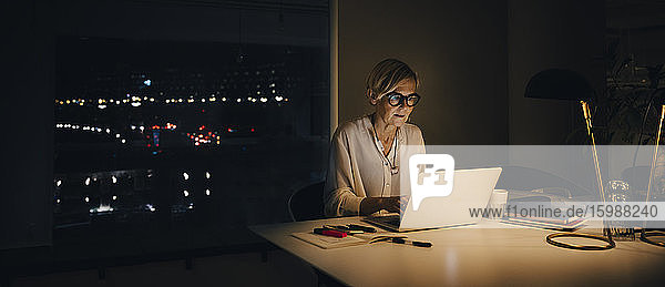 Reife Berufstätige  die spät arbeiten  während sie ihren Laptop am beleuchteten Schreibtisch im Büro benutzen