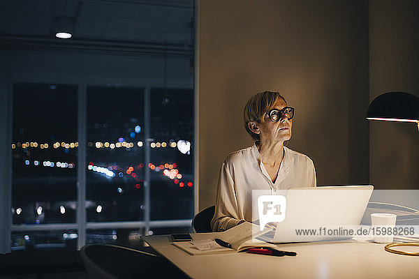 Nachdenkliche Geschäftsfrau sitzt mit Laptop am beleuchteten Schreibtisch im Kreativbüro