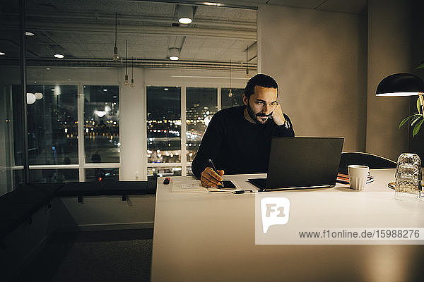 Müde Geschäftsmann arbeitet spät  während er am Schreibtisch im beleuchteten Kreativbüro sitzt
