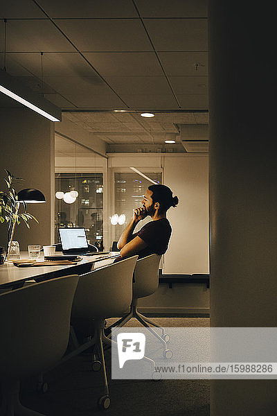 Seitenansicht eines nachdenklichen Geschäftsmannes  der spät arbeitet  während er am kreativen Arbeitsplatz auf einen Laptop schaut