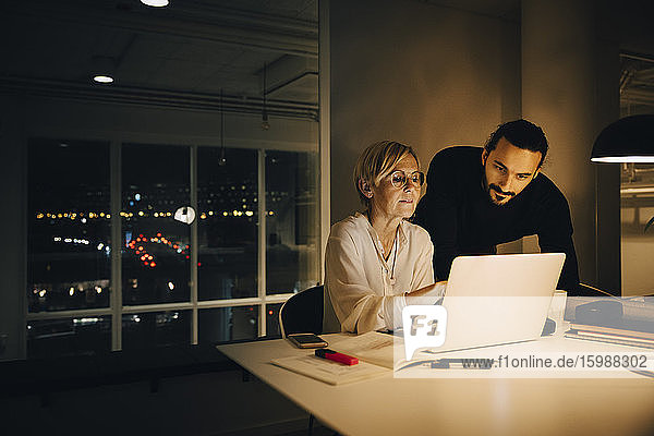 Selbstbewusste reife Geschäftsfrau diskutiert am Laptop mit Geschäftsmann am beleuchteten Schreibtisch im Kreativbüro