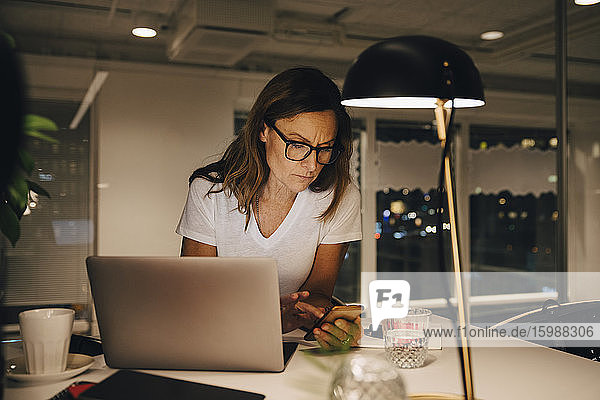 Selbstbewusste Unternehmerin  die ein Smartphone benutzt  während sie nachts mit ihrem Laptop an einem beleuchteten Schreibtisch am Arbeitsplatz sitzt