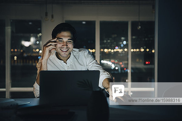 Lächelnder männlicher Profi arbeitet spät  während er in einem dunklen Coworking Space auf einen Laptop schaut