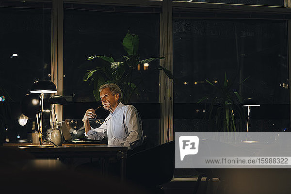 Thoughtful senior male professional looking away while sitting at desk in dark office