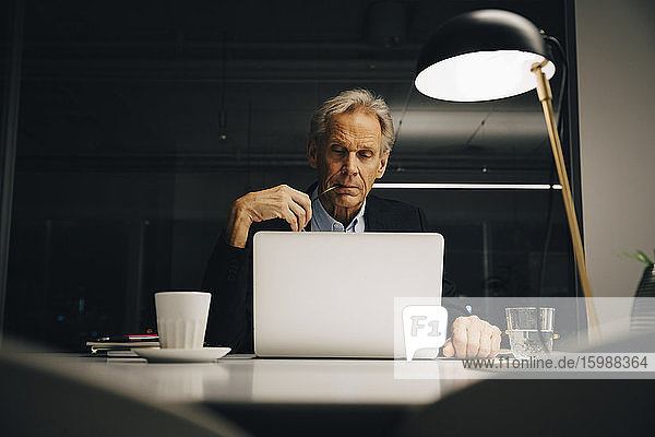 Engagierter älterer männlicher Unternehmer schaut auf den Laptop  während er bis spät in der kreativen Arbeitswelt arbeitet