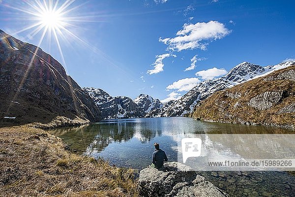 Young man sitting on a rock  mountains reflected in the lake  Lake Harris  Conical Hill  Routeburn Track  Mount Aspiring National Park  Westland District  West Coast  South Island  New Zealand  Oceania