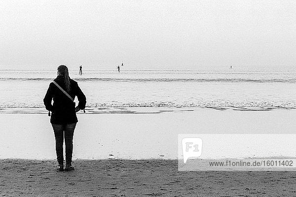 Scheveningen  Niederlande. Junge erwachsene  kaukasische Frau  die kurz vor Sonnenuntergang vom Strand aus über die Nordsee starrt und schaut. Aufgenommen auf analogem Kodak T-Max Schwarz-Weiß-Film im Jahr 2019.