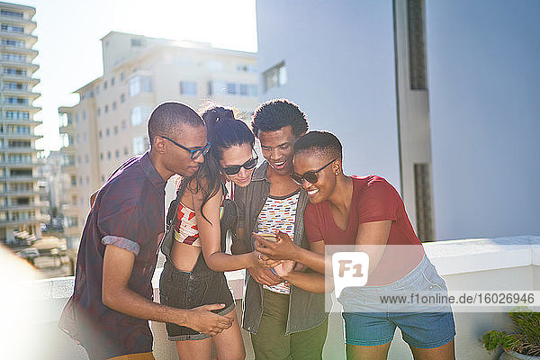Young friends using smart phone on sunny rooftop balcony