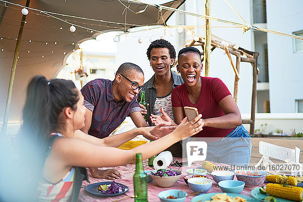 Glückliche junge Freunde  die ein Smartphone benutzen und am Balkontisch essen