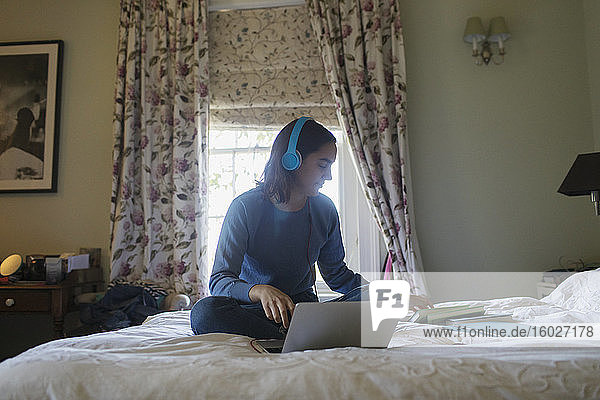 Teenage girl with headphones and laptop studying on bed