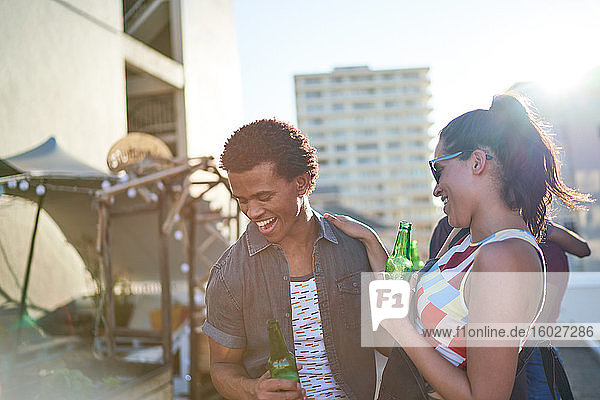 Happy young couple drinking beer on sunny urban rooftop balcony