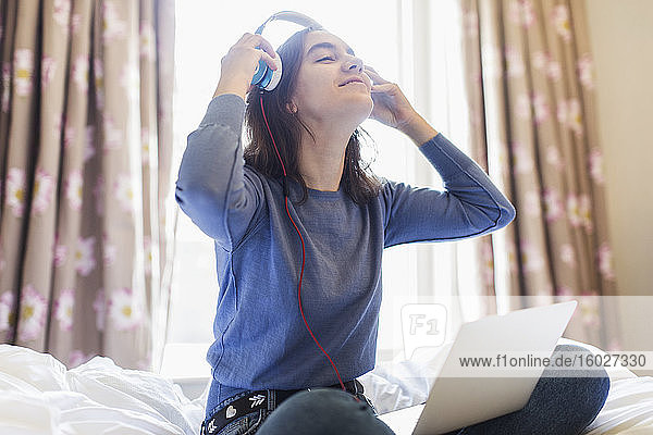 Lächelndes Teenager-Mädchen mit Kopfhörern und Laptop auf sonnigem Bett