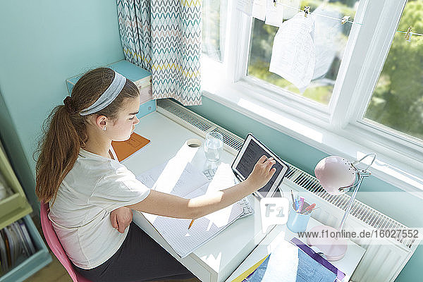 Mädchen mit digitalem Tablet beim Heimunterricht am Schreibtisch im sonnigen Schlafzimmer