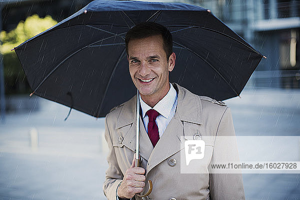 Lächelnder Geschäftsmann im Trenchcoat unterm Regenschirm
