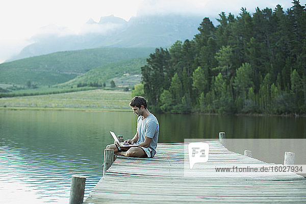Man using laptop on dock over calm lake