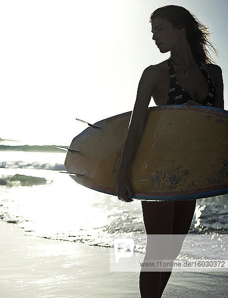 Frau  die an einem Sandstrand am Meer steht und ein Surfbrett in der Hand hält.