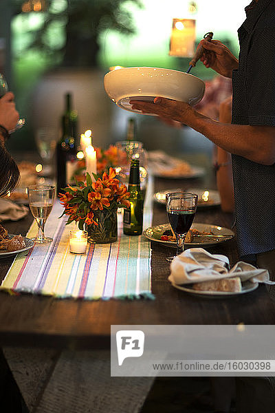 Person  die an einem Tisch steht und eine Schüssel  Weingläser  Teller  Blumen und Kerzen auf dem Tisch hält.