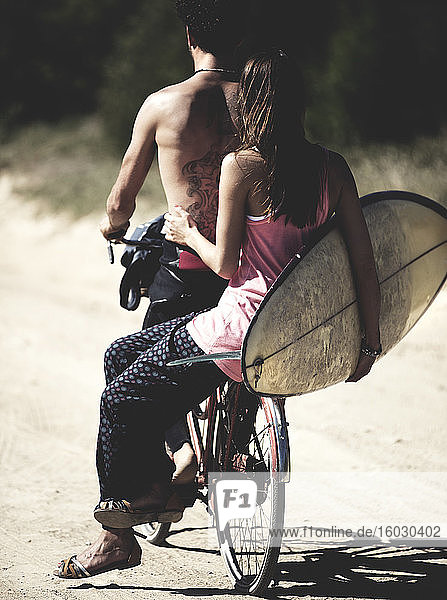 Rückansicht von Mann und Frau auf einem Fahrrad  Frau sitzt auf einem Gepäckträger und hält ein Surfbrett.