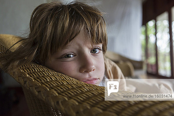 Ein müder fünfjähriger Junge  der sich in einem Hotel in einem Korbstuhl ausruht und in die Kamera schaut.