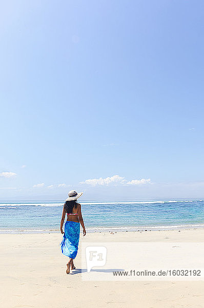 Eine junge Frau mit Sonnenhut und Schal  die an einem unberührten tropischen Strand auf der Insel Bali spazieren geht  Indonesien  Südostasien  Asien