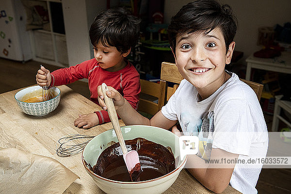 Two boys with black hair sitting at a kitchen table  baking chocolate cake.