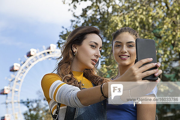 Two young women with long brown hair standing in a park near a Ferris wheel  taking selfie with mobile phone.