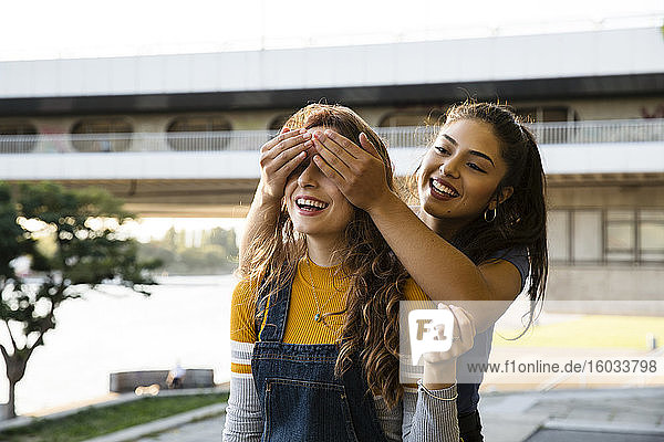 Zwei junge Frauen mit langen braunen Haaren stehen am Flussufer  lächelnd und die Augen bedeckend.