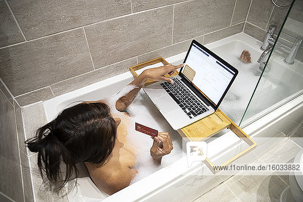 Frau sitzt in der Badewanne  nimmt ein Schaumbad und kauft während der Coronavirus-Krise online auf ihrem Laptop ein.
