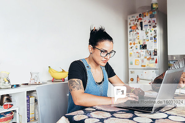 Frau mit langen braunen Haaren mit Brille  die an einem Küchentisch sitzt und einen Laptop benutzt.