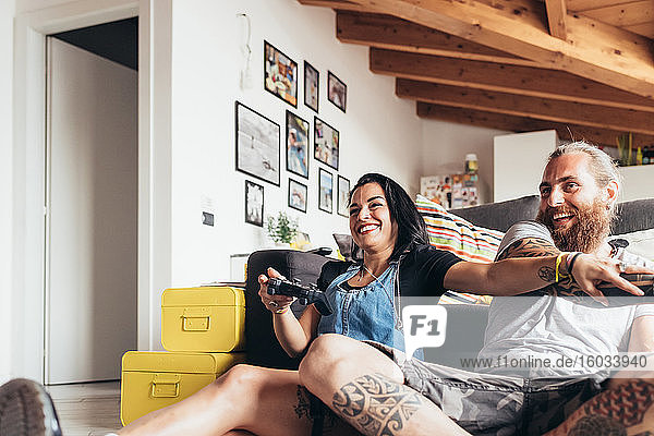 Bearded tattooed man with long brunette hair and woman with long brown hair sitting on a sofa  smiling while playing console game.