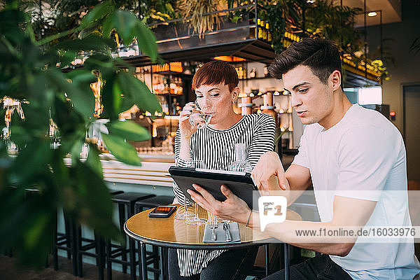 Junge Frau und Mann sitzen an einem Tisch in einer Bar und schauen auf ein digitales Tablett.