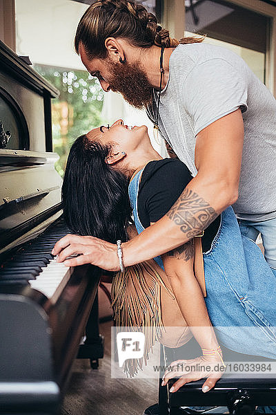 Bearded tattooed man with long brunette hair leaning over smiling woman  playing piano.