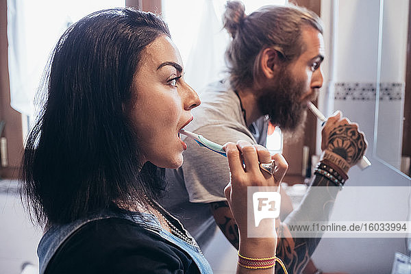 Bearded tattooed man with long brunette hair and woman with long brown hair standing in front of mirror  brushing their teeth.